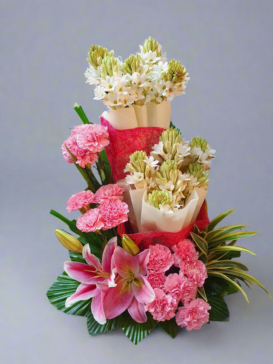 Discover the beauty of delicate pink carnations, vibrant pink lilies, lush green leaves, and fragrant tuberoses in a stunning floral arrangement.